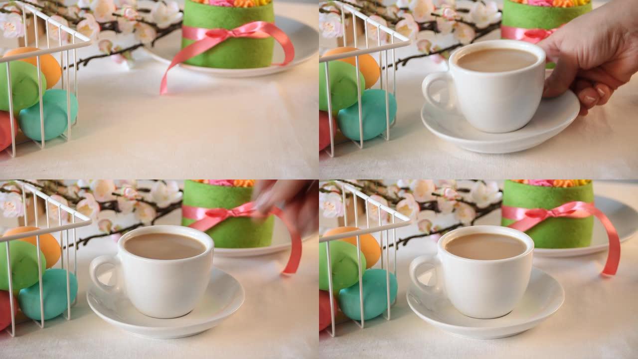 春季复活节节日餐桌。绿色小蛋糕，背景上有黄油奶油花和彩色鸡蛋。女人的手在桌子上放一杯茶或咖啡