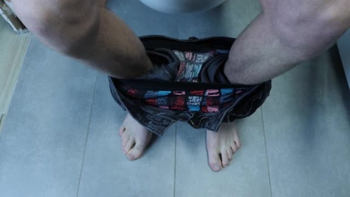 男人坐在马桶上，放下卫生纸，特写镜头。穿着牛仔裤的男性腿的俯视图，赤脚坐在灰色浴室的马桶上。