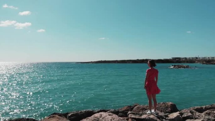 穿着红色连衣裙的迷人女人站在地中海的岩石悬崖上，看着蓝色的海水和天空。站在塞浦路斯帕福斯珊瑚湾岩石码