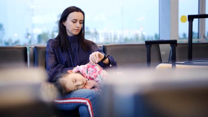 一位母亲抱着她的小女儿，在等待下一个航班时睡在机场航站楼的椅子上