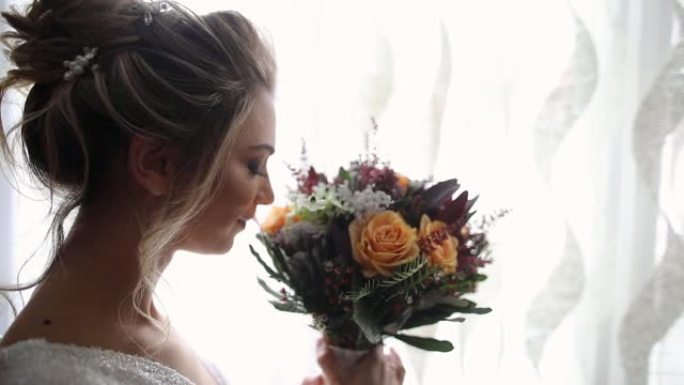 美丽的年轻女子站在窗户附近，拿着并闻到华丽的婚礼花束