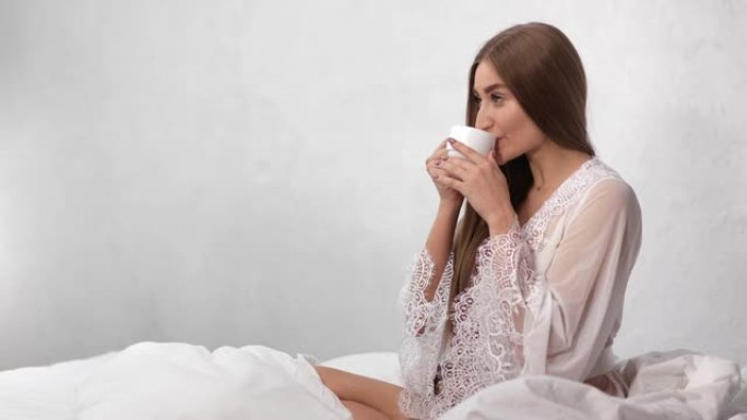 穿着性感睡衣的微笑晨女孩在床上喝咖啡。红色相机上的中等镜头