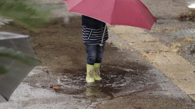 一个男孩在雨中站在水坑里，拿着雨伞和橡胶靴。