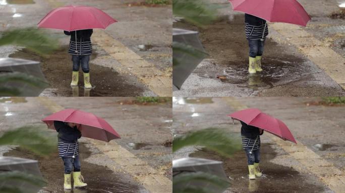 一个男孩在雨中站在水坑里，拿着雨伞和橡胶靴。
