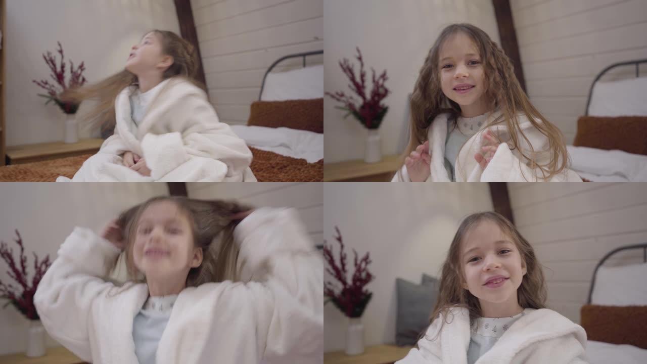相机接近可爱的白色浴袍高加索小女孩，摇着长长的卷发。洗完澡后坐在床上微笑的漂亮孩子。休闲，生活方式，