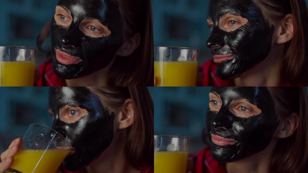 30多岁的年轻女子脸上戴着黑色化妆面具，看着相机喝一杯橙汁。蓝色厨房的女人美容程序。特写