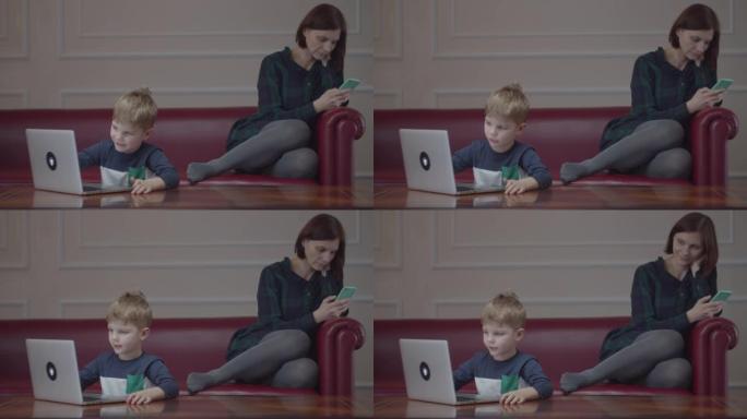 金发碧眼的学龄前男孩在笔记本电脑上打字，微笑着看着电脑屏幕。妈妈拿着手机坐在沙发上。孩子坐在桌旁在家
