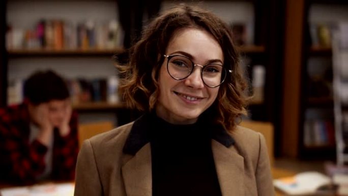 一个戴着眼镜的迷人短发欧洲女学生站在高中图书馆微笑着看着相机微笑的肖像。教育、文学和人民观念。背景模