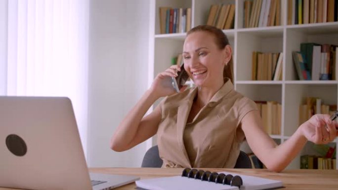 开朗的女商人在笔记本电脑前谈论手机，在办公室里轻松地玩眼镜。