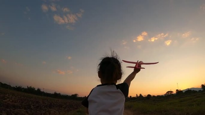 一个快乐的小女孩的慢动作在日落背景下与玩具飞机一起奔跑。幸福家庭和童年梦想的概念。