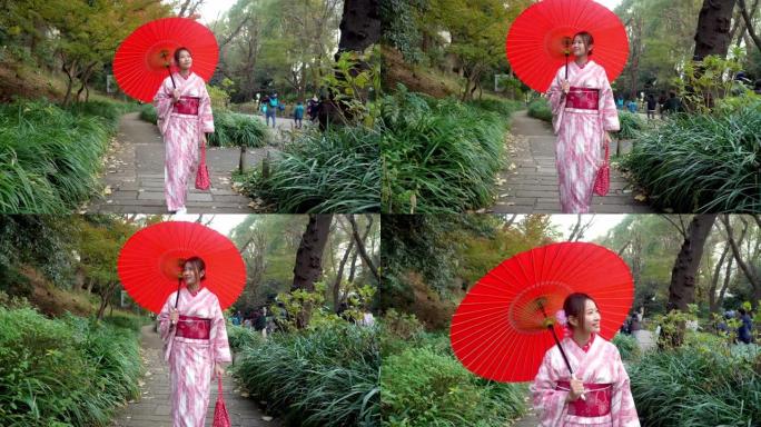 4k快乐年轻美丽的亚洲女性女孩穿着粉色传统日本和服连衣裙，拿着红色的雨伞，走在公园的街道上，看着远处
