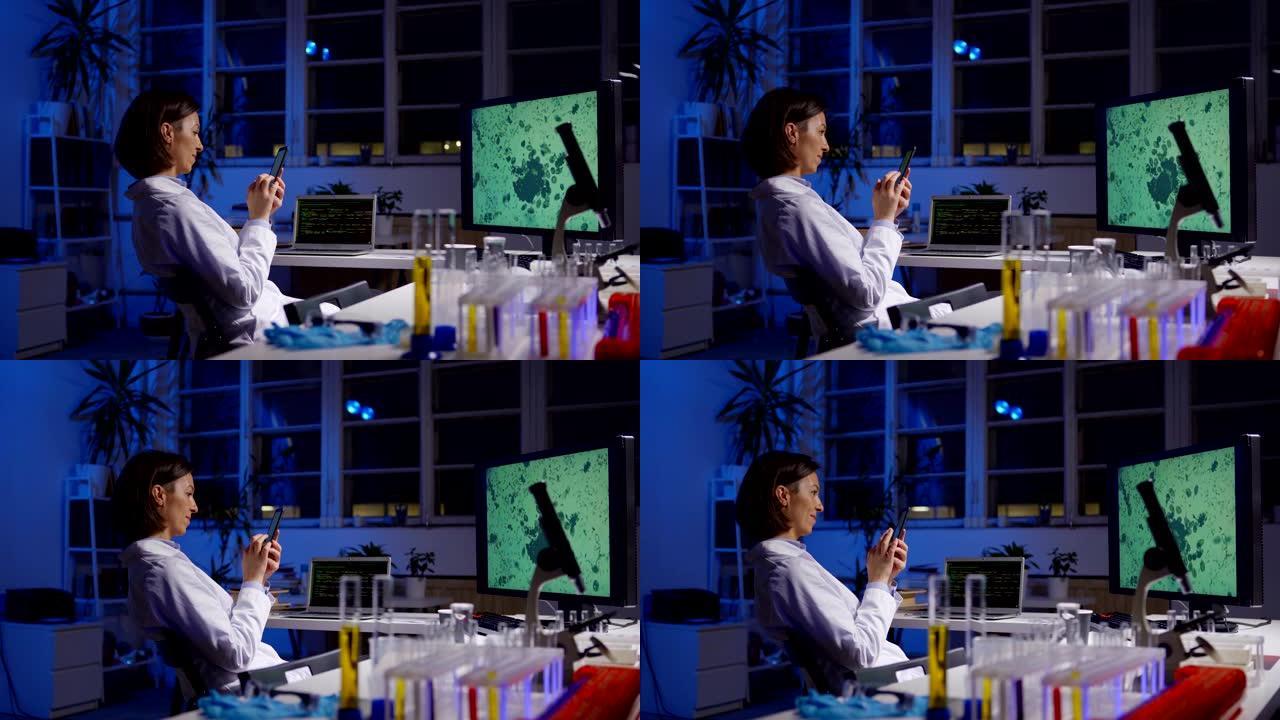 穿着实验室外套浏览智能手机的中年女科学家的侧视图跟踪镜头坐在实验室的桌子上，放大了她的计算机屏幕上的