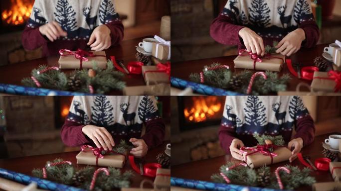 近距离观察人的双手在壁炉附近的圣诞节假期礼物上系弓。男性用纸包装礼物盒，用杉木树枝，圆锥体，木桌上的