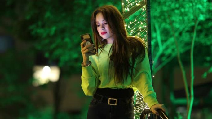 坦率美丽的女孩在晚上使用智能手机。20多岁的千禧一代年轻女子晚上看着屏幕