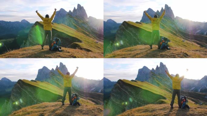 年轻人在Seceda山上徒步旅行，在日出时向山顶举起手臂。背包，黄色夹克，靴子，豆豆。前往意大利特伦