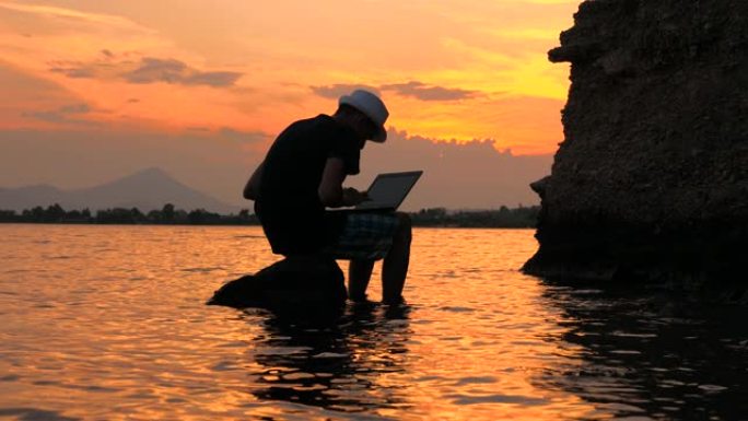 快乐的人坐在石头上，在电脑上工作，看到海景，静态框架。日落海景背景上的男性剪影。在海边工作的旅游博客