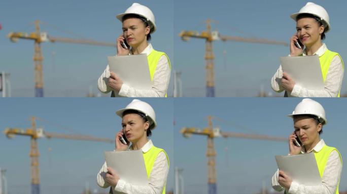 戴着头盔的建筑工人拿着一个装有文件的文件夹，并在电话上交谈。成功的商业女性