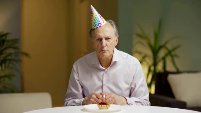 沮丧的戴礼帽的老人坐在公寓的桌子旁，看着蜡烛燃烧的小生日蛋糕，然后抬起头，不高兴地看着相机
