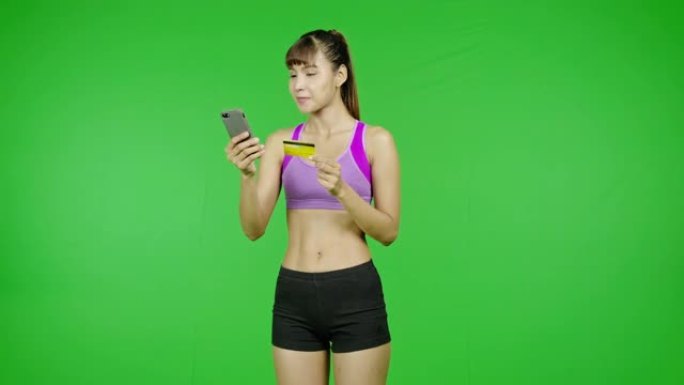 Fit女人用手机刷卡购买，绿屏背景，Fit身材，网上购买，惊喜心情。亚洲女运动员穿着紫色的衣服站在镜