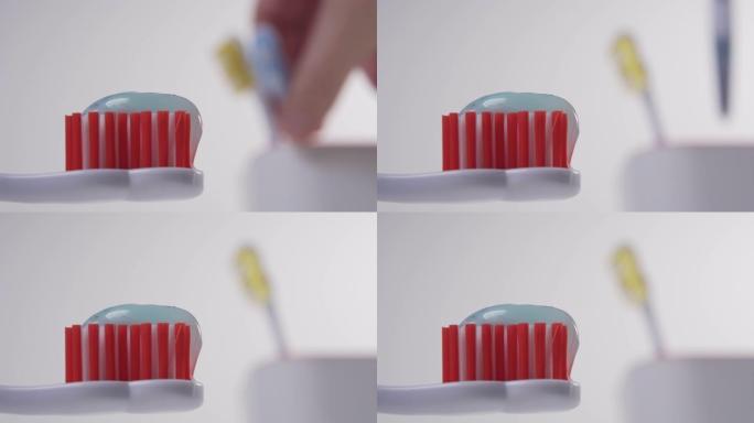 白色牙刷，红色刷毛，顶部有蓝色牙膏。在背景中，一只手从玻璃杯中拿起蓝色牙刷
