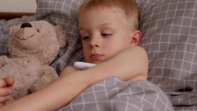 一个生病的小男孩躺在床上，体温计和泰迪熊的特写镜头。