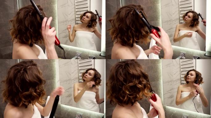 一个穿着白色大浴巾的年轻女子在镜子前用卷发棒烫头发。一个女孩洗完澡后在浴室里做发型。透过镜子观察。近