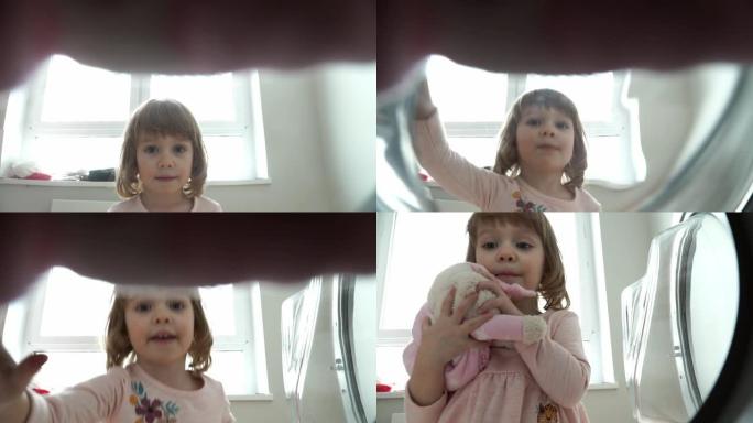 3岁女孩的肖像特写打开洗衣机的舱门，洗涤后拿出泰迪熊玩具，拥抱他并微笑。Amera看着孩子的洗衣机
