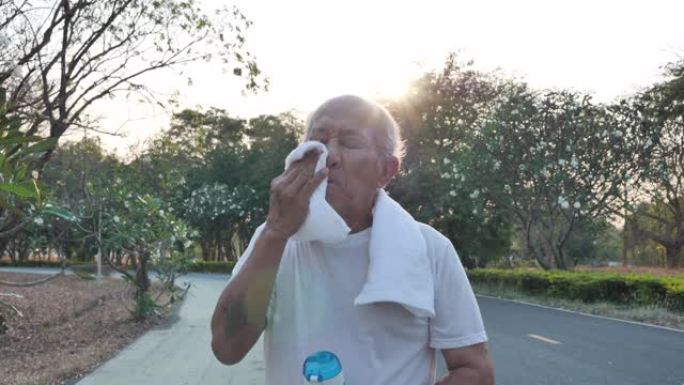 亚洲老人在晚上有阳光的公园里跑步后，用毛巾擦汗。健康生活方式和运动理念。慢动作