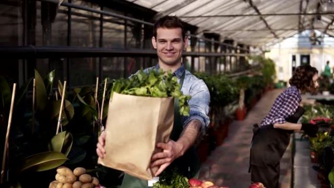穿着围裙的年轻高加索高个子男人拿着新鲜蔬菜和蔬菜的棕色纸袋，微笑着笑着，在食品市场上摆姿势拍照，站在