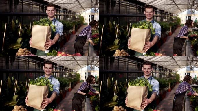 穿着围裙的年轻高加索高个子男人拿着新鲜蔬菜和蔬菜的棕色纸袋，微笑着笑着，在食品市场上摆姿势拍照，站在