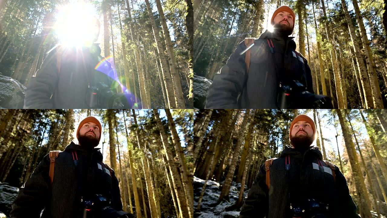 一位认真的大胡子摄影师的肖像，背着背包和相机站在针叶树的冬季森林中，在背光和阳光照射下