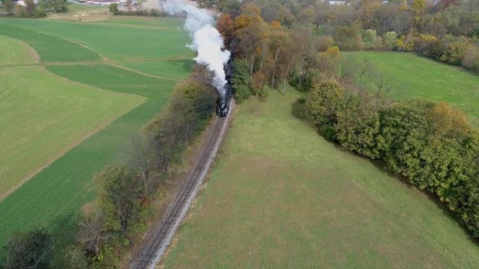 一辆经过修复的古董蒸汽机车在吹着白烟和蒸汽的过程中穿越秋天的树木的空中俯视