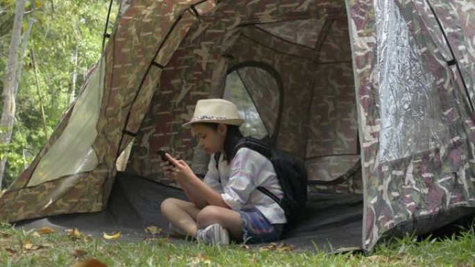 亚洲可爱女孩，背包坐着，在自然公园的帐篷前使用手机。