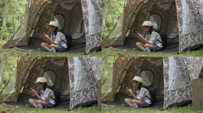 亚洲可爱女孩，背包坐着，在自然公园的帐篷前使用手机。