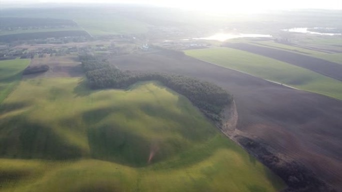 空中无人机镜头: 飞越农业田和山丘上郁郁葱葱的树叶绿色景观，被日落照亮。