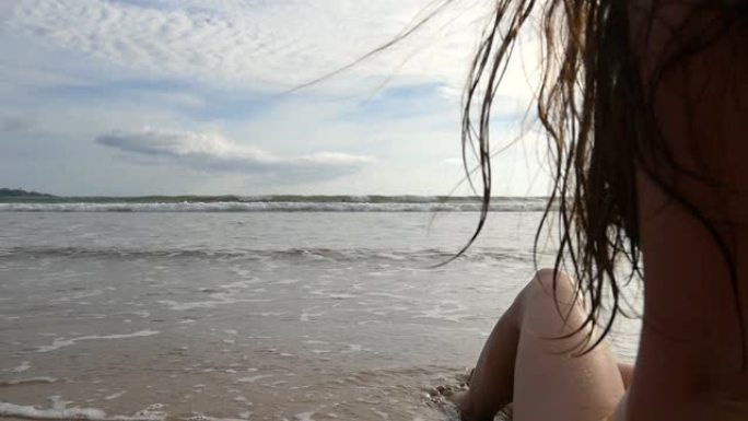 穿着比基尼的年轻女子坐在海边，晴天晒日光浴。海浪在海岸上冲刷着年轻的女性脚。背景上美丽的海景。暑假或
