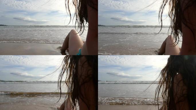 穿着比基尼的年轻女子坐在海边，晴天晒日光浴。海浪在海岸上冲刷着年轻的女性脚。背景上美丽的海景。暑假或