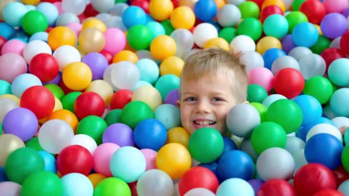 金发小男孩躺在游戏中心大干戏水池里的多色塑料球上。对着镜头微笑。肖像特写。在游戏室玩得开心。休闲活动