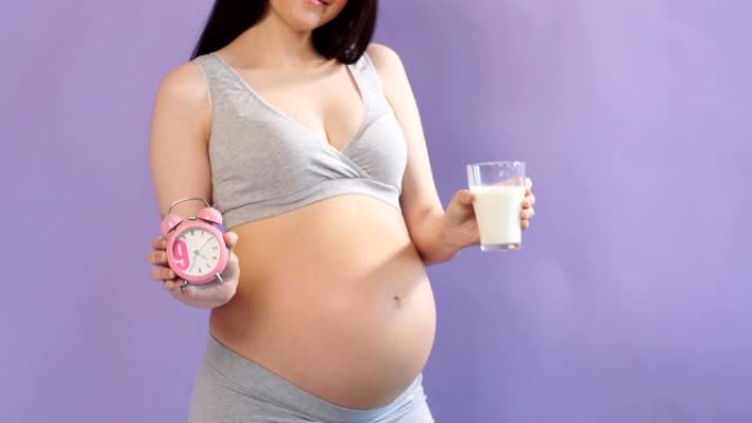 年轻的怀孕白种人模型，留着一头黑发，拿着一杯牛奶和一个闹钟