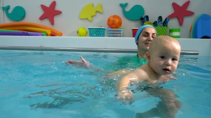 母亲帮助婴儿在儿童游泳池游泳，并用双手在水中支撑婴儿。他们玩耍和大笑。游泳训练。婴儿游泳。特写