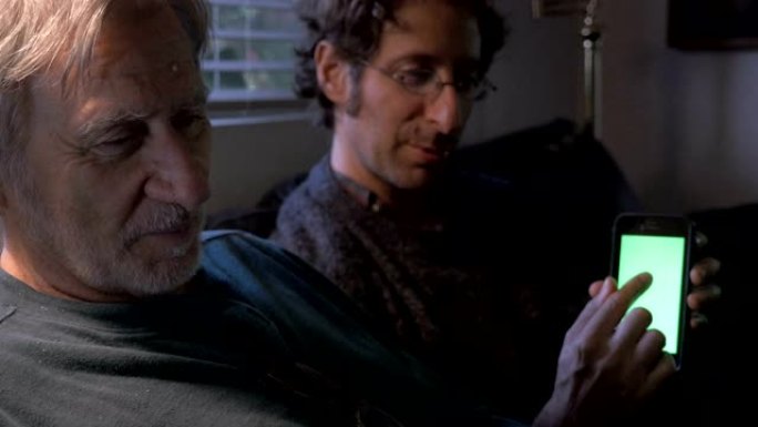 一位上了年纪的父亲正在操作平板电脑，而他的小儿子正在向他展示自己的手机