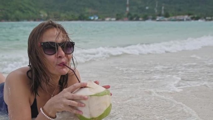 一名年轻女子躺在沙滩上。这个女孩正在喝椰子水。椰子鸡尾酒。热带岛屿上微笑的女人。波浪中的女孩。