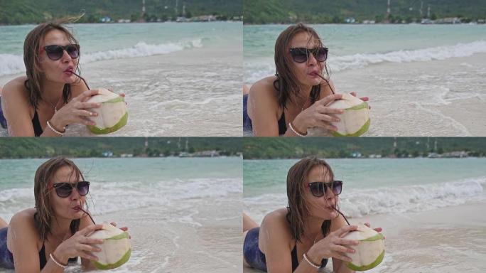 一名年轻女子躺在沙滩上。这个女孩正在喝椰子水。椰子鸡尾酒。热带岛屿上微笑的女人。波浪中的女孩。