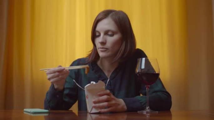 30多岁的年轻女性成年人在家用手机用筷子吃亚洲盒饭，喝葡萄酒。女人独自拿着手机和葡萄酒。