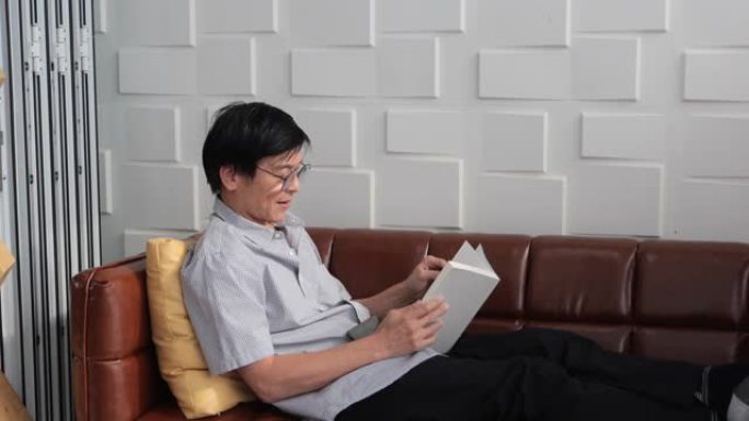 亚洲老人在家客厅沙发上看书，亚洲老人与阅读杂志一起放松和快乐