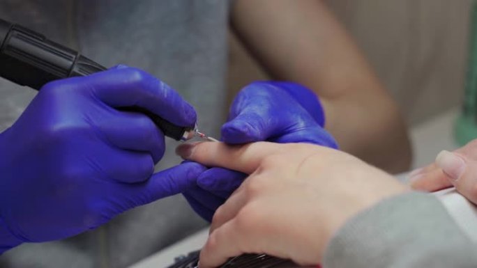 美容院戴蓝色手套的女性修指甲大师使用修指甲机处理客户指甲上的角质层