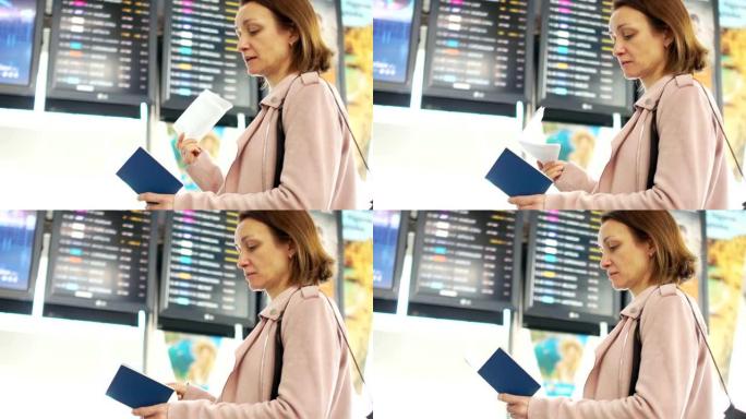 成熟的女人独自站在机场航班时刻表附近。查看出发，飞机到达机场的信息板，并在机票上选择并与出口号码进行