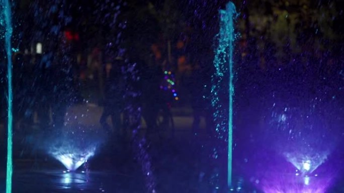 晚上五颜六色的舞蹈喷泉。黑暗背景上的水流的慢动作特写镜头。无法识别的人剪影