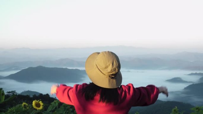 亚洲女性旅行者坐在岩石上，看着日出山景，举起手臂呼吸新鲜空气