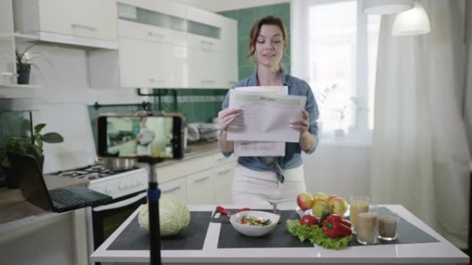有吸引力的女孩在智能手机上为追随者写vlog视频，告诉他们在餐桌上直播时在相机上的饮食计划节目，厨房